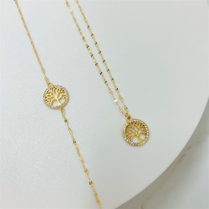 Könnte beinhalten: Zwei goldfarbene Halsketten mit Lebensbaum-Anhängern. Eine Halskette hat eine längere Kette mit einem kreisförmigen Anhänger, die andere eine kürzere Kette. Die Anhänger sind mit kleinen, klaren Kristallen verziert.