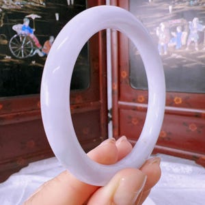 May include: A smooth, solid lavender-colored bangle bracelet with a rounded profile. The bangle has a polished, semi-translucent appearance and a consistent circular shape, showcasing a soft purple hue throughout the entire piece of jewelry.