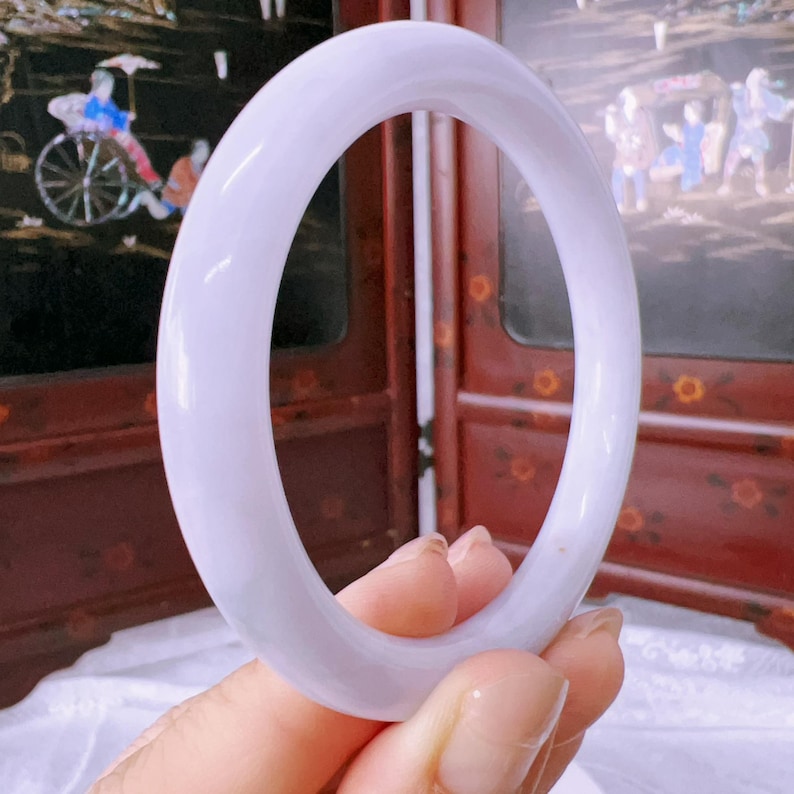May include: A smooth, solid lavender-colored bangle bracelet with a rounded profile. The bangle has a polished, semi-translucent appearance and a consistent circular shape, showcasing a soft purple hue throughout the entire piece of jewelry.