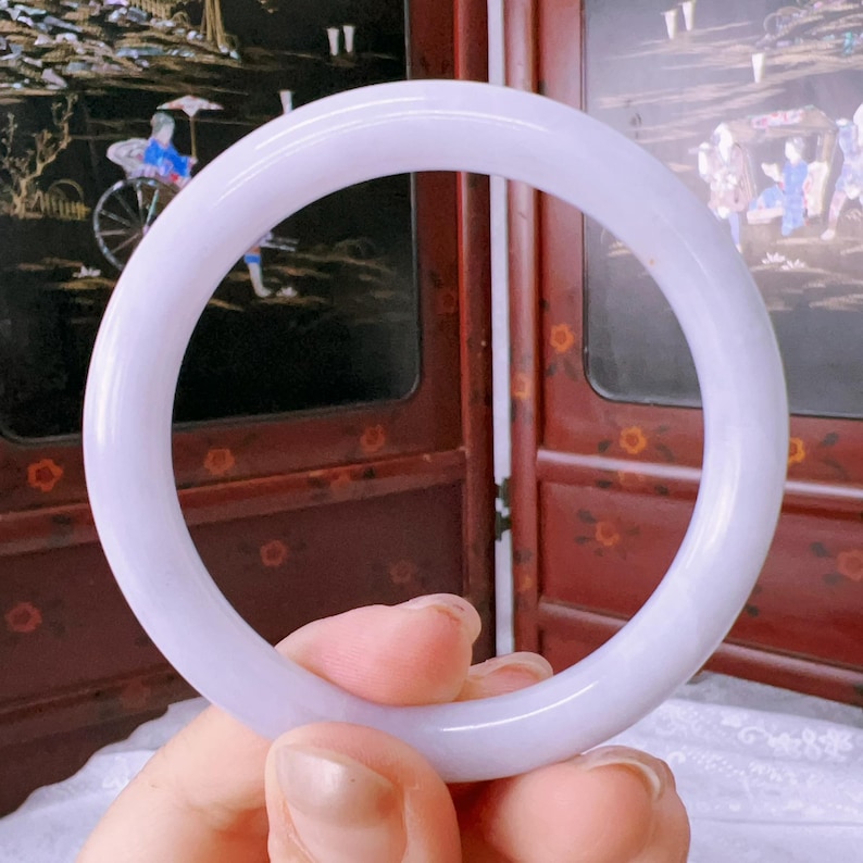 May include: A smooth, circular bangle bracelet crafted from translucent lavender-colored stone. The bangle features a polished, rounded surface with a consistent thickness throughout its entire circumference, showcasing a soft, uniform light purple hue.