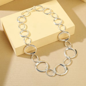 May include: A silver-toned necklace featuring a series of interconnected oval and circular links. The necklace is displayed on a light yellow surface, showcasing its reflective qualities. The design is modern and elegant, suitable for various occasions.