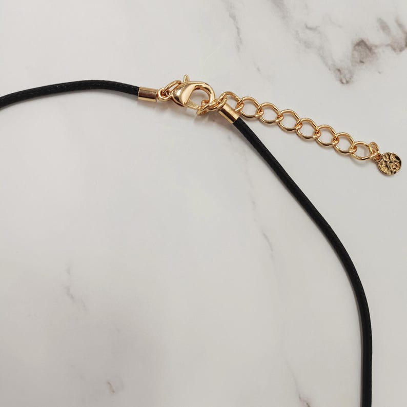 May include: Close-up of a black cord necklace with gold-coloured clasp and extender chain. The clasp is a lobster claw style, and the extender chain has a small charm at the end. The background is a white marble pattern.