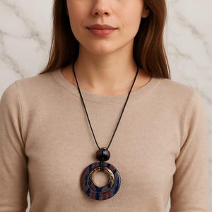 May include: A statement necklace with a large, layered pendant. The pendant features a gold-coloured inner ring and a dark blue and red marbled pattern. The necklace hangs from a black cord, creating a bold contrast.