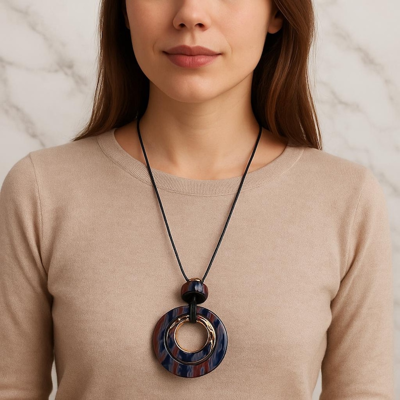 May include: A statement necklace with a large, layered pendant. The pendant features a gold-coloured inner ring and a dark blue and red marbled pattern. The necklace hangs from a black cord, creating a bold contrast.