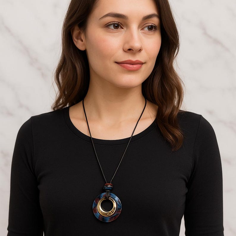 May include: A long necklace with a circular pendant in marbled blue, red, and gold. The pendant hangs from a black cord. The woman in the image is wearing a black long-sleeved top. The necklace is a statement piece.