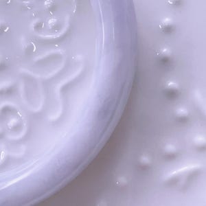 May include: A white ceramic plate featuring a raised, embossed floral pattern on the rim and a textured surface with small, evenly spaced raised dots across the center. The glossy finish reflects light, highlighting the intricate relief details.