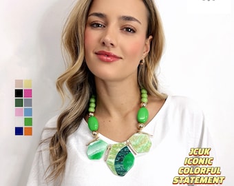 Op de afbeelding: Een vrouw draagt een wit shirt met een groene en gouden statementketting. De ketting heeft grote, geometrische groene en witte kralen. De tekst "JCUk ICONIC COLORFUL STATEMENT NECKLACE" is zichtbaar in de rechterbenedenhoek van de afbeelding.