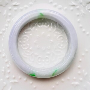May include: A smooth, circular bangle bracelet crafted from translucent lavender jadeite with distinct, vibrant green inclusions. The bangle features a polished, rounded surface and a consistent thickness, showcasing natural colour variations throughout the stone.