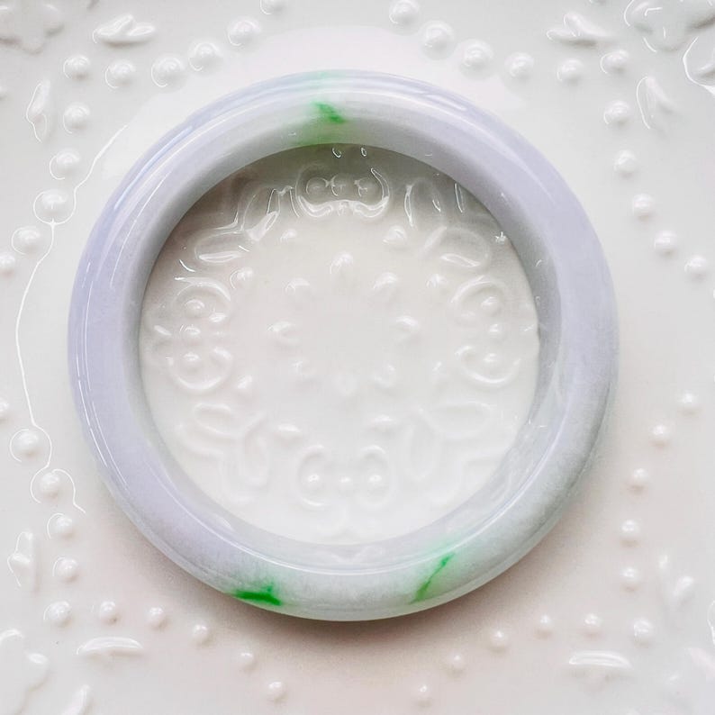 May include: A smooth, circular bangle bracelet crafted from translucent lavender jadeite with distinct, vibrant green inclusions. The bangle features a polished, rounded surface and a consistent thickness, showcasing natural colour variations throughout the stone.