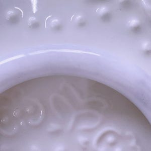 May include: A white ceramic object featuring a smooth, curved rim and a textured surface with raised, rounded dots and a subtle, embossed floral pattern. The material has a glossy finish that reflects light, highlighting the intricate details.