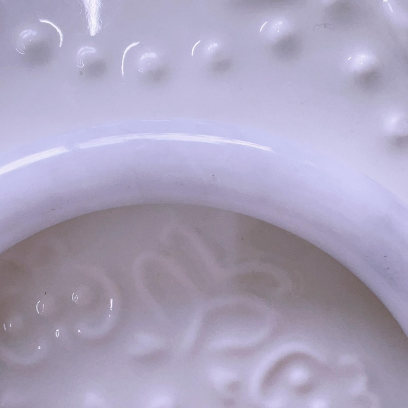May include: A white ceramic object featuring a smooth, curved rim and a textured surface with raised, rounded dots and a subtle, embossed floral pattern. The material has a glossy finish that reflects light, highlighting the intricate details.