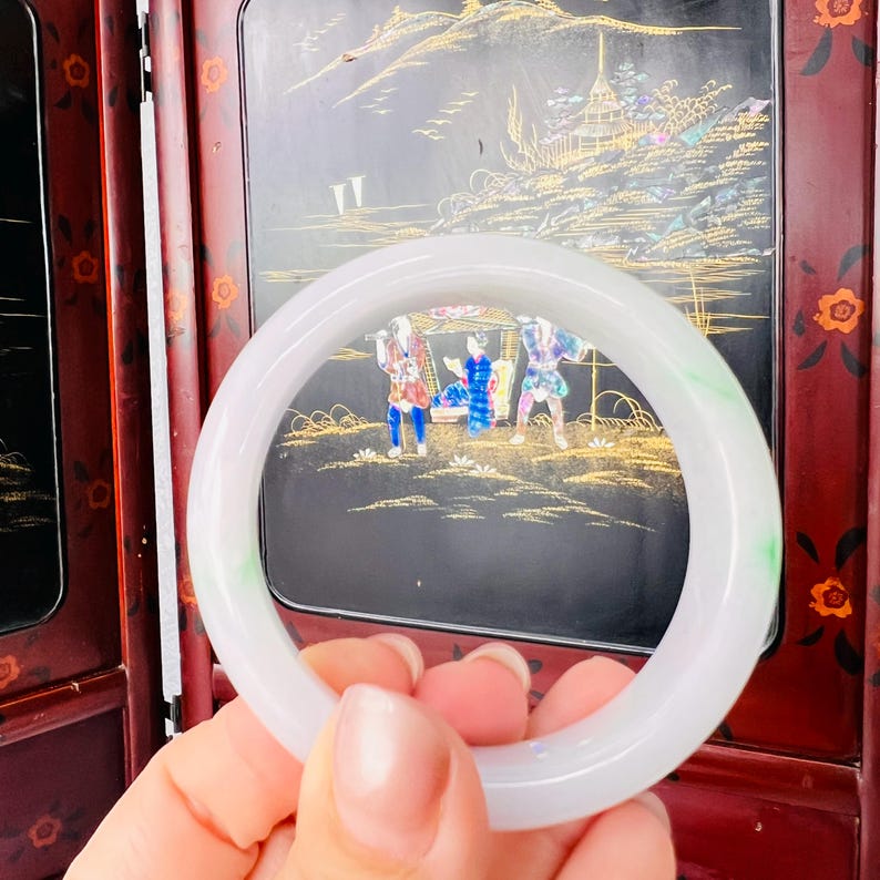 May include: A smooth, translucent white jade bangle bracelet featuring subtle, faint green inclusions throughout the circular band. The polished stone surface reflects light, showcasing a consistent, rounded profile and a solid, continuous structure.