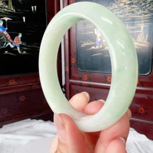 May include: A smooth, polished bangle bracelet crafted from pale, translucent light green jade. The circular jewelry piece features a rounded profile with a consistent thickness and a soft, glossy finish that catches the light across its surface.