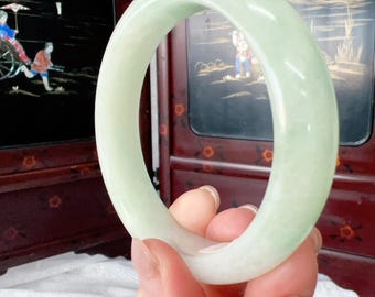 May include: A smooth, polished bangle bracelet crafted from pale, translucent light green jade. The circular jewellery piece features a rounded profile with a consistent thickness and a soft, glossy finish that catches the light across its surface.