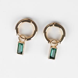 Pode incluir: Um par de brincos de argola dourados com um pendente retangular de pedra preciosa verde esmeralda. Os brincos são exibidos sobre um fundo branco, realçando o design e a cor da joia.