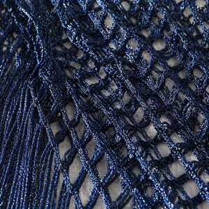 May include: Close-up of a navy blue, open-weave fabric with a shimmering, metallic finish. The netting-like material has a textured appearance, with long, fringed strands cascading down the left side. The fabric's intricate design is highlighted by the play of light.