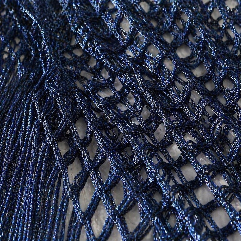 May include: Close-up of a navy blue, open-weave fabric with a shimmering, metallic finish. The netting-like material has a textured appearance, with long, fringed strands cascading down the left side. The fabric's intricate design is highlighted by the play of light.