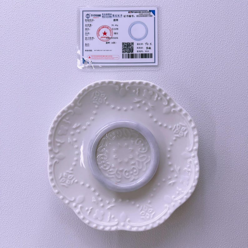 May include: A smooth, light lavender-grey jade bangle bracelet resting on a white ceramic saucer with an embossed floral pattern. Above the saucer is a rectangular white certification card featuring Chinese text, a red seal, and a barcode.