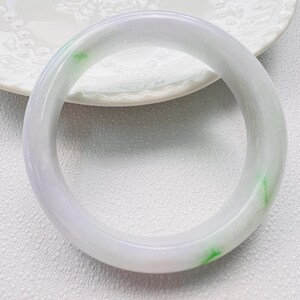May include: A smooth, polished bangle bracelet crafted from translucent white stone with subtle lavender undertones and distinct, vibrant green inclusions scattered throughout the circular band, featuring a rounded profile and a glossy, reflective surface.