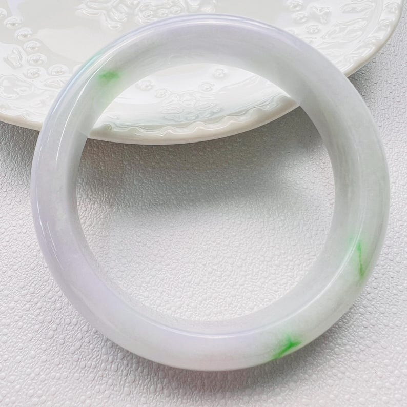May include: A smooth, polished bangle bracelet crafted from translucent white stone with subtle lavender undertones and distinct, vibrant green inclusions scattered throughout the circular band, featuring a rounded profile and a glossy, reflective surface.