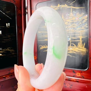 May include: A smooth, polished bangle bracelet crafted from translucent white jadeite with distinct, vibrant green inclusions. The circular accessory features a rounded, substantial profile with a glossy, reflective surface that catches light.