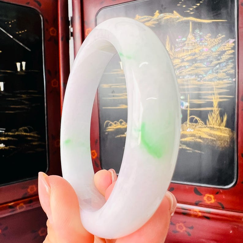 May include: A smooth, polished bangle bracelet crafted from translucent white jadeite with distinct, vibrant green inclusions. The circular accessory features a rounded, substantial profile with a glossy, reflective surface that catches light.