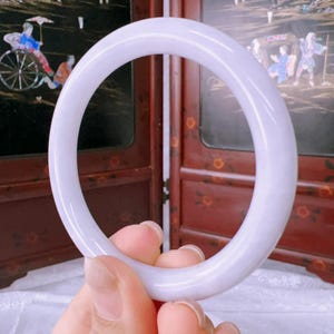 May include: A smooth, circular bangle bracelet crafted from solid lavender-colored stone. The bangle features a polished, glossy finish with a consistent, rounded profile throughout its entire circumference, showcasing a soft, light purple hue.