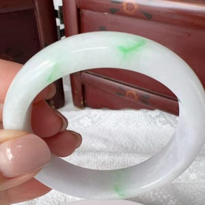 May include: A smooth, polished bangle bracelet crafted from translucent white jadeite stone, featuring subtle, natural streaks of vibrant green colour throughout the circular band, showcasing a high-gloss finish and a rounded, solid construction.