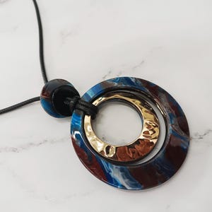 May include: A statement pendant necklace with a marbled blue and brown circular pendant and a gold-coloured inner ring. The necklace has a black cord and a matching marbled bead. The pendant is a striking piece of jewellery.