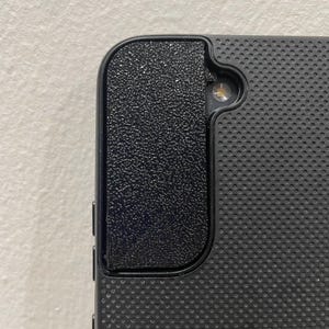 Puede incluir: Funda de teléfono negra con un patrón texturizado y punteado. La funda tiene una sección lateral texturizada en relieve y un recorte para la cámara. La funda está diseñada para proteger el teléfono.