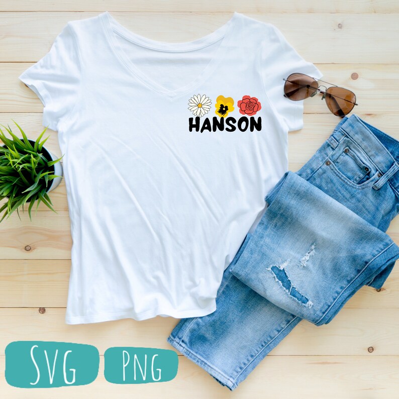 May include: White t-shirt with a graphic of three flowers and the word "HANSON" in black text. The t-shirt is laying on a wooden surface with a pair of blue jeans and sunglasses.