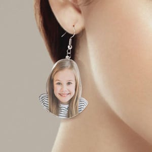 May include: A custom dangle earring featuring a flat, die-cut acrylic pendant shaped like a portrait of a smiling child with long blonde hair, wearing a white and black striped shirt. The pendant hangs from a silver-toned metal fishhook wire.