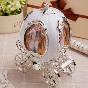 May include: A white and silver carriage-shaped photo frame. The frame has two oval openings for photos and decorative silver wheels. The carriage is decorated with ornate silver trim and a floral design on top. The photos show a woman and child.