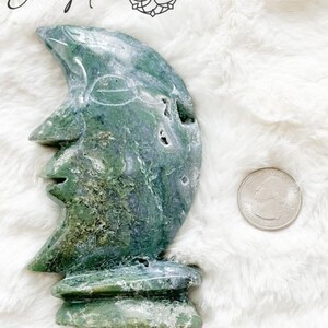 May include: Green and white carved moonstone sculpture with a face on the front. The sculpture is sitting on a white fluffy surface. View 2.