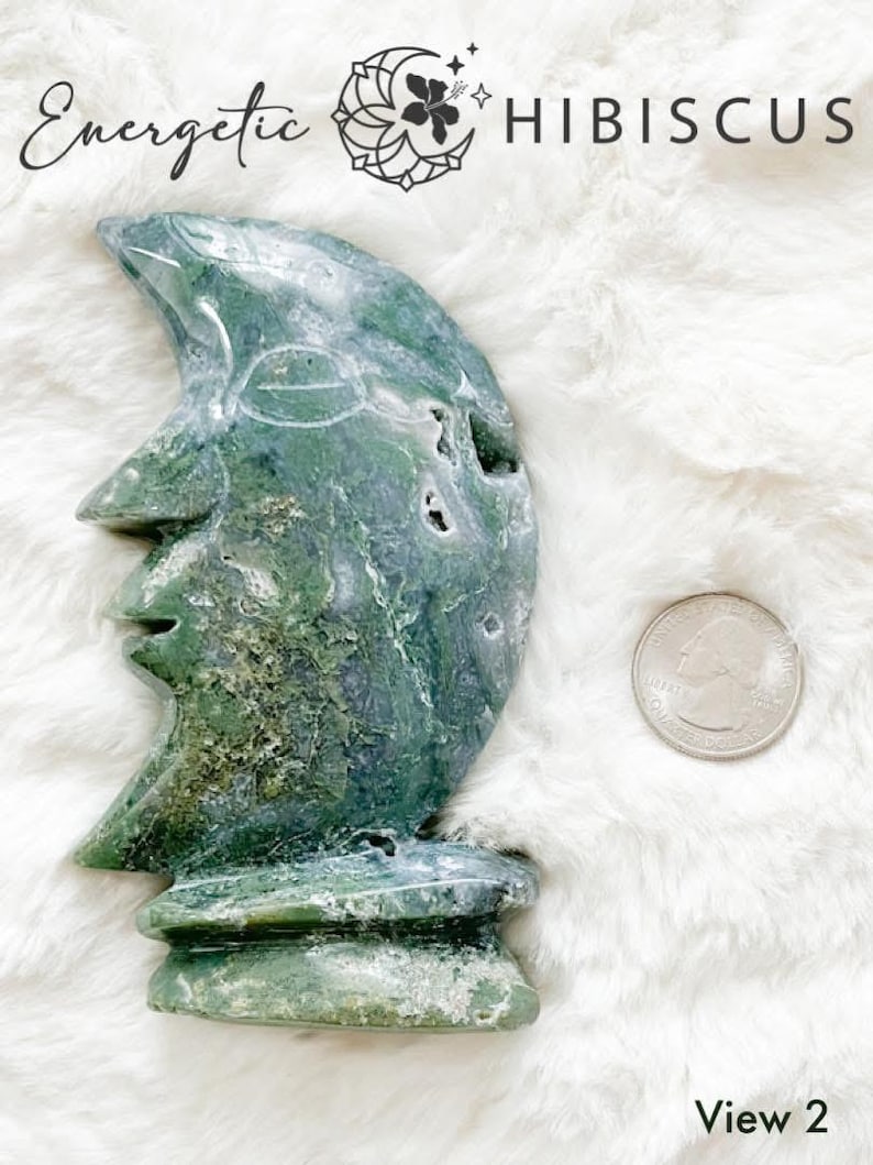 May include: Green and white carved moonstone sculpture with a face on the front. The sculpture is sitting on a white fluffy surface. View 2.