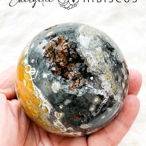 May include: A polished, spherical stone with a geode center. The stone features a mix of colors, including black, gray, yellow, and brown. The geode center is filled with sparkling crystals. The text "Energetic Hibiscus" is at the top. Approximate size: 70mm.
