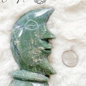 May include: Green and white moon-shaped stone sculpture, carved with a face, standing on a white fluffy surface. Approx: 5" tall.