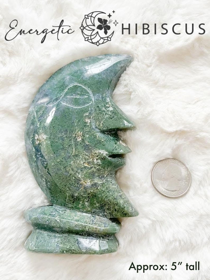 May include: Green and white moon-shaped stone sculpture, carved with a face, standing on a white fluffy surface. Approx: 5" tall.