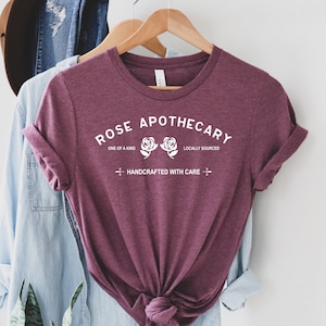 May include: A maroon t-shirt with the text "ROSE APOTHECARY" in white. The text is surrounded by two white roses and the words "ONE OF A KIND" and "LOCALLY SOURCED". Below the roses is the text "HANDCRAFTED WITH CARE".