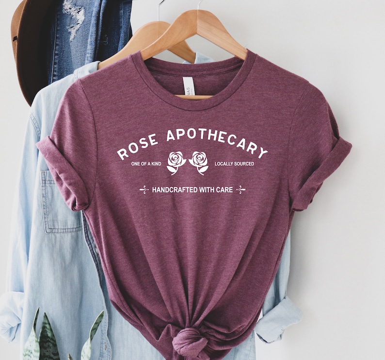 May include: A maroon t-shirt with the text "ROSE APOTHECARY" in white. The text is surrounded by two white roses and the words "ONE OF A KIND" and "LOCALLY SOURCED". Below the roses is the text "HANDCRAFTED WITH CARE".