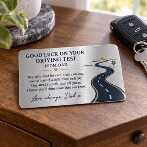 Peut inclure: Une carte portefeuille rectangulaire en métal brossé avec le texte GOOD LUCK ON YOUR DRIVING TEST FROM DAD, un poème, et un graphique représentant une route sinueuse menant vers un soleil à l'horizon avec une petite icône de trèfle vert.