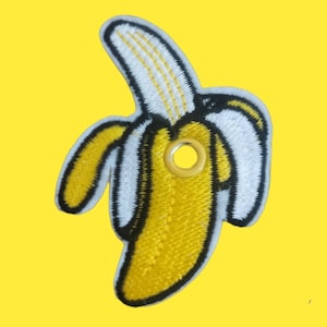 May include: A yellow embroidered banana patch with a black outline and a white stem. The patch has a metal eyelet for attaching it to a fabric item.