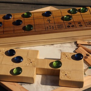 May include: A wooden board game with a light wood base and a darker wood top. The game includes blue and green glass marbles, wooden game pieces, and decorative carvings. The game is displayed on a wooden surface.