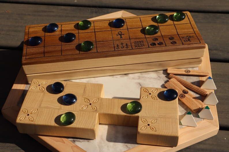 May include: A wooden board game with a light wood base and a darker wood top. The game includes blue and green glass marbles, wooden game pieces, and decorative carvings. The game is displayed on a wooden surface.