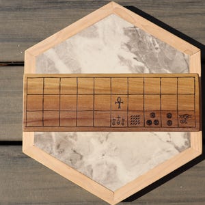 May include: A wooden board game with a light wood frame and a marble-like center. The game board features a grid pattern and engraved symbols. The overall design is geometric and the game is set on a wooden surface.