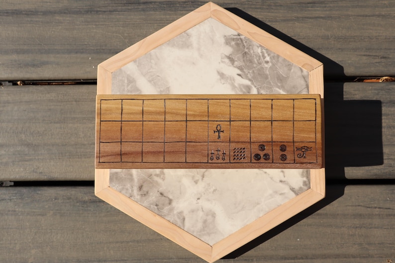 May include: A wooden board game with a light wood frame and a marble-like center. The game board features a grid pattern and engraved symbols. The overall design is geometric and the game is set on a wooden surface.