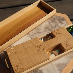 May include: Wooden game box with a light brown finish. The box is open, revealing a compartment with green and blue glass marbles. The lid is separate, and the box has a decorative floral design.