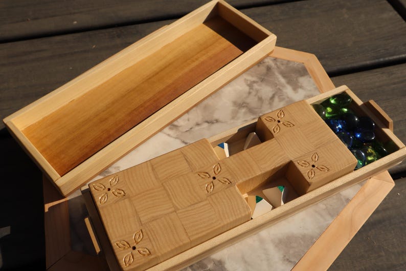 May include: Wooden game box with a light brown finish. The box is open, revealing a compartment with green and blue glass marbles. The lid is separate, and the box has a decorative floral design.