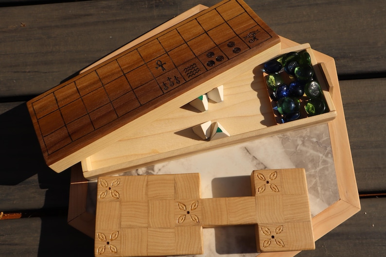 May include: Wooden board game set with a light wood finish. The set includes a rectangular game board with a grid pattern, a tray of glass marbles, and other wooden game pieces. The game board has etched symbols.