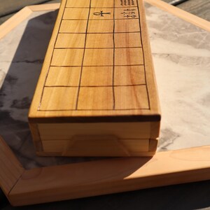 May include: A light-colored wooden box with a grid pattern on the top surface. The box has an ankh symbol and other hieroglyphic-like symbols. The box is on a marble-patterned surface.